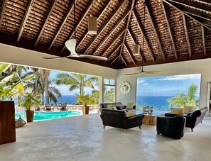 A spacious living area with a large window overlooking a pool and the ocean, featuring a thatched ceiling and modern furniture. - Tropical Hilltop Luxury Private Villa with Pool including Butler. (Montego Bay)