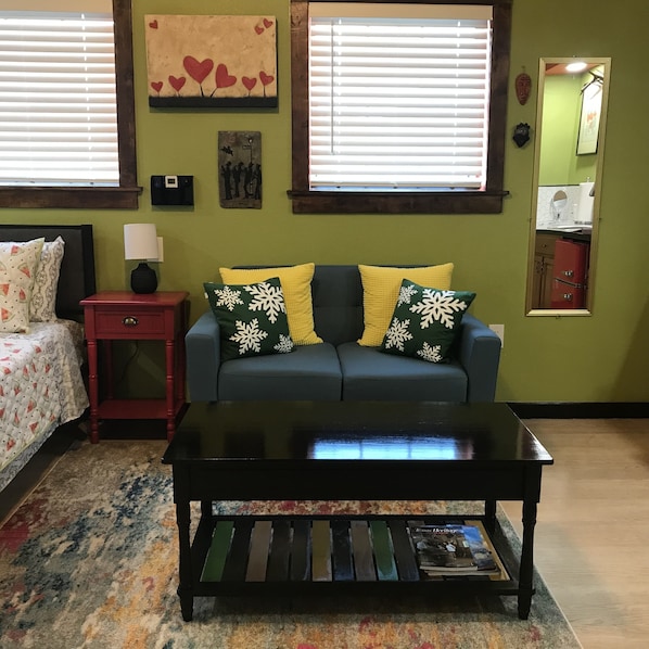 Una sala de estar con un sofá azul, almohadas amarillas y verdes, una mesita de noche roja, una mesa de centro negra y una alfombra estampada.
