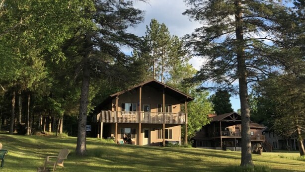 Eine Lodge mit einer großen Terrasse und zahlreichen Fenstern, umgeben von hohen Bäumen und einer Rasenfläche.