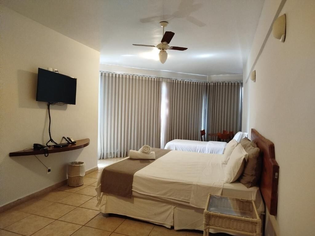 A hotel room with a bed, a television, a ceiling fan, and a window with curtains.