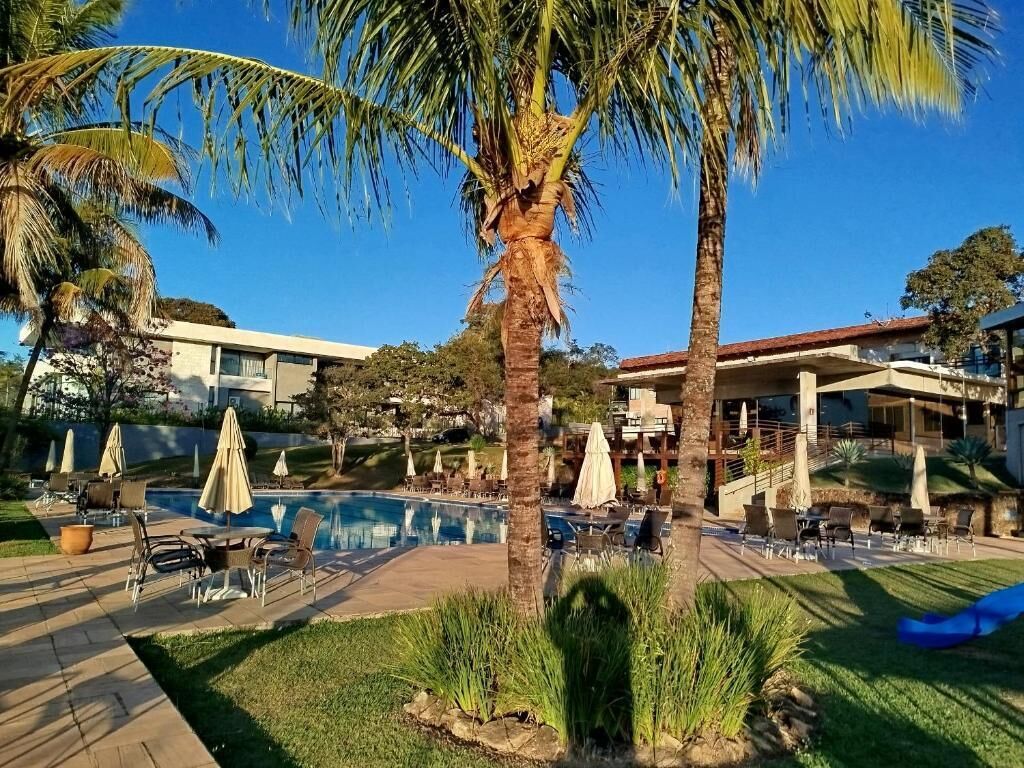 Un área de piscina con sillas de descanso y sombrillas, rodeada de palmeras y un edificio con techo rojo.