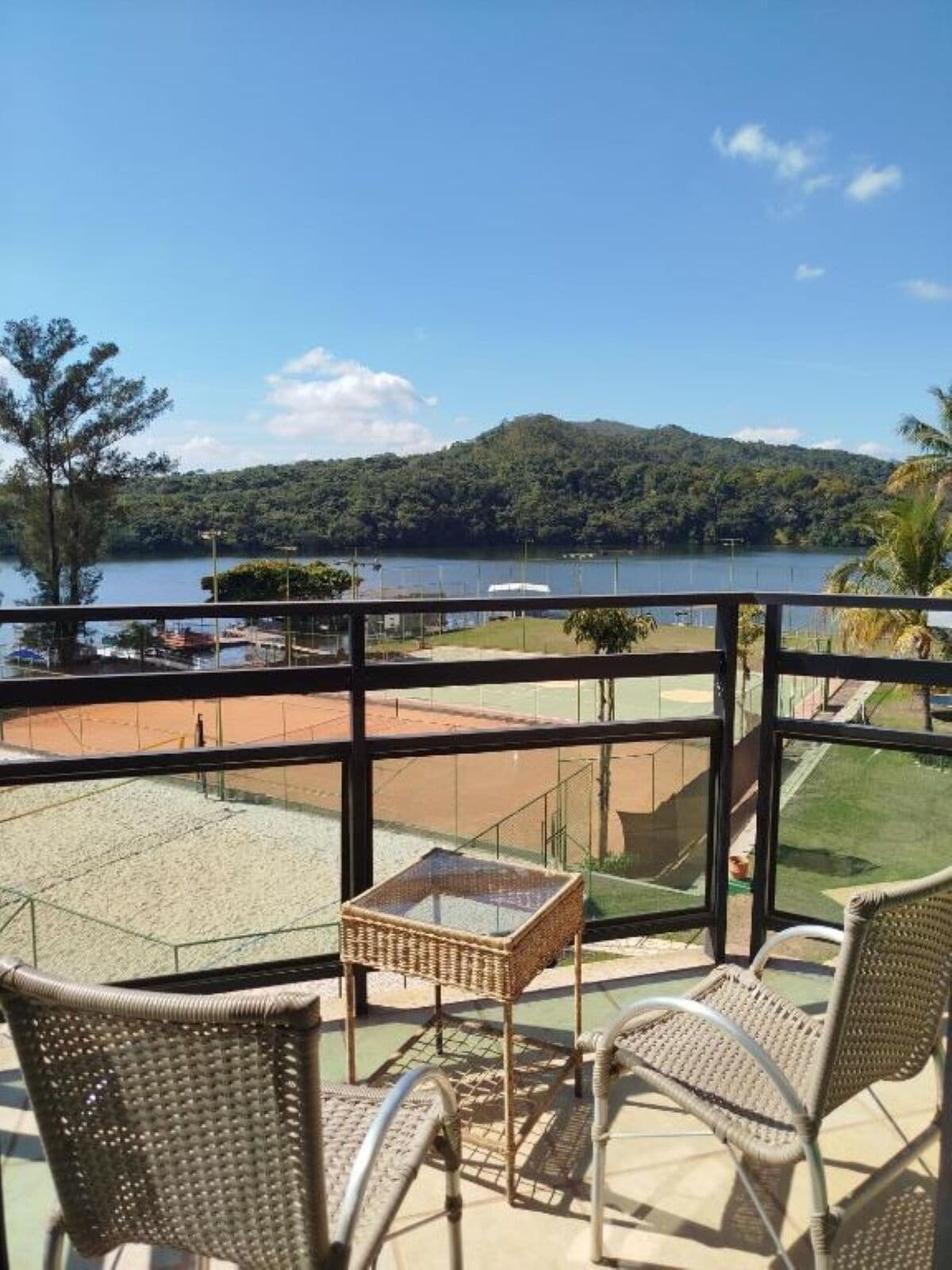 Un balcón con sillas de mimbre y una pequeña mesa, con vista a un lago y una cancha de tenis.