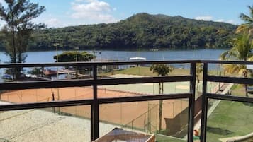 Un balcón con sillas de mimbre y una pequeña mesa, con vista a un lago y una cancha de tenis.