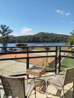 Un balcón con sillas de mimbre y una pequeña mesa, con vista a un lago y una cancha de tenis.
