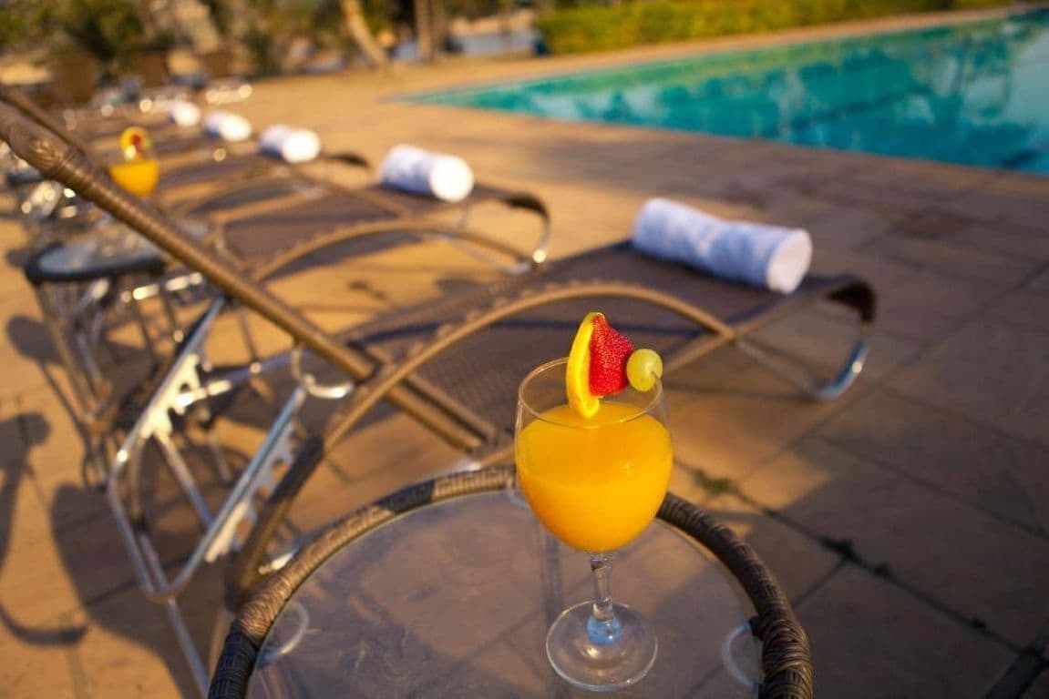A glass of orange juice with a slice of fruit on a glass table by a poolside lounge chair.