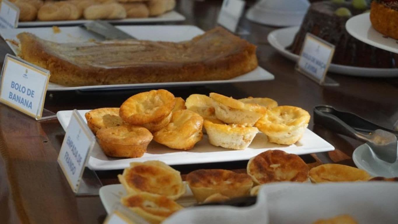 Variedad de productos horneados a la vista, incluyendo un letrero que dice 'Bolo de Banana' y 'Empanadas de Frango'.