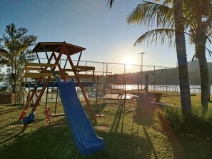 Un parque infantil con tobogán y columpios, rodeado de césped y palmeras, con una masa de agua y un cielo despejado al fondo.