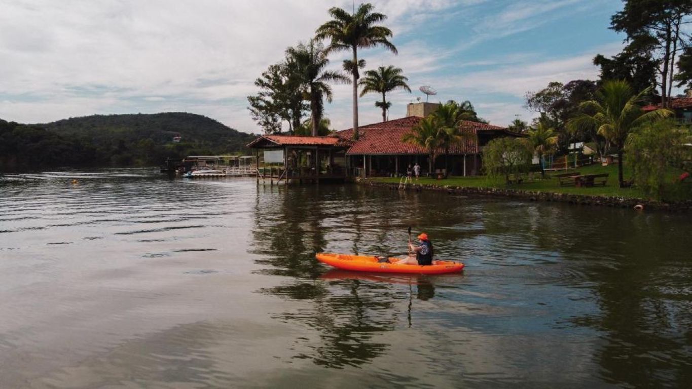 Un lago tranquilo con un kayakista en un kayak naranja, rodeado de un paisaje tropical con palmeras y un edificio de techo rojo.
