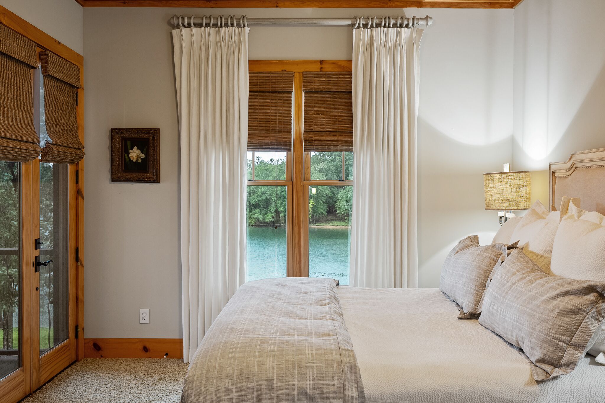 A bedroom with a large bed, a window with curtains, a bedside lamp, and a view of a lake.