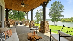 An outdoor patio with a wooden ceiling, stone pillars, and a view of a lake. There are seating areas with cushions and a table with a bottle and glasses. - Luxurious French Country Estate on Smith Lake- Flat yard, dock, fiber internet! (Arley)