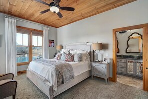 A bedroom with a bed, bedside tables, a ceiling fan, a window with curtains, and a view of the outside.