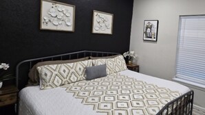 Un dormitorio con cama, una mesita de noche, una pared decorada con cuadros y una ventana con persianas.