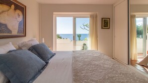 Une chambre avec un grand lit, un tableau accroché au mur et une vue sur la mer par la fenêtre.
