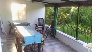 Een overdekt terras met een tafel en stoelen, een raam met gordijnen en uitzicht op groen.