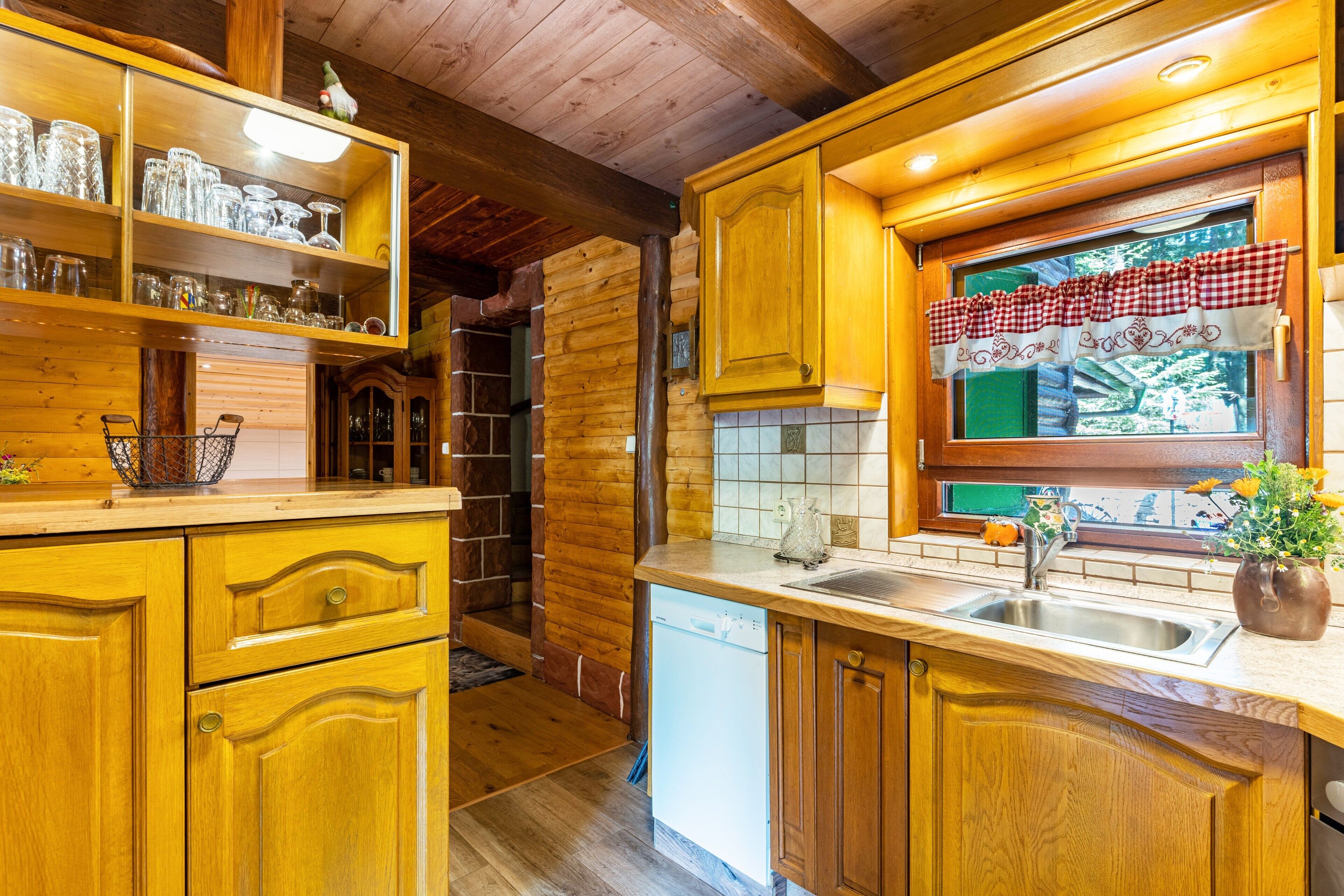A wooden interior with a kitchen area, including a sink, cabinets, and a window with a checkered curtain.