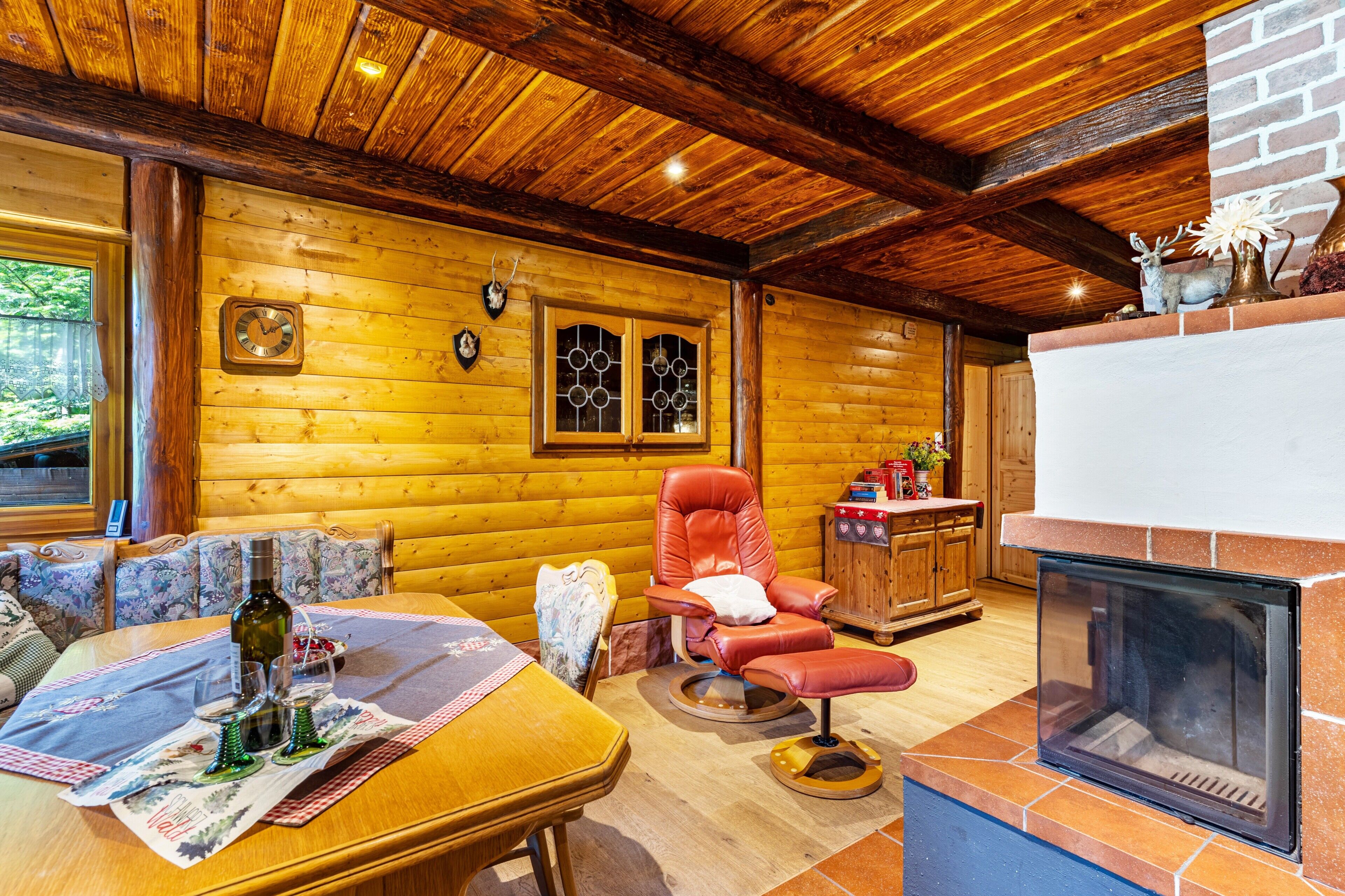 Interior de una cabaña de madera con chimenea, un sillón de cuero, una mesa de comedor con copas de vino y un reloj en la pared.