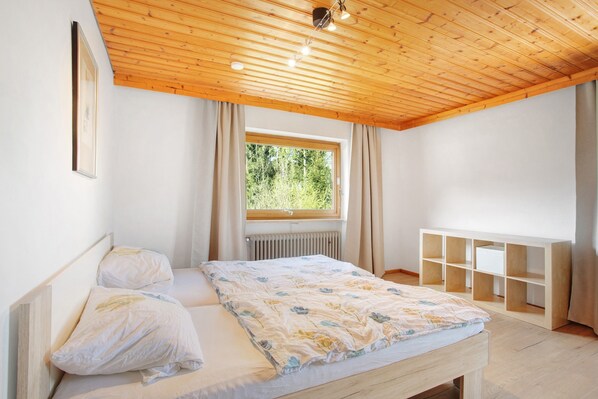 A bedroom with a wooden ceiling, a bed with floral bedding, a window with curtains, and a built-in shelving unit.