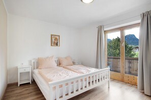 A bedroom with a wooden floor, a white bed with a headboard, a nightstand, a window with curtains, and a view of greenery and mountains.