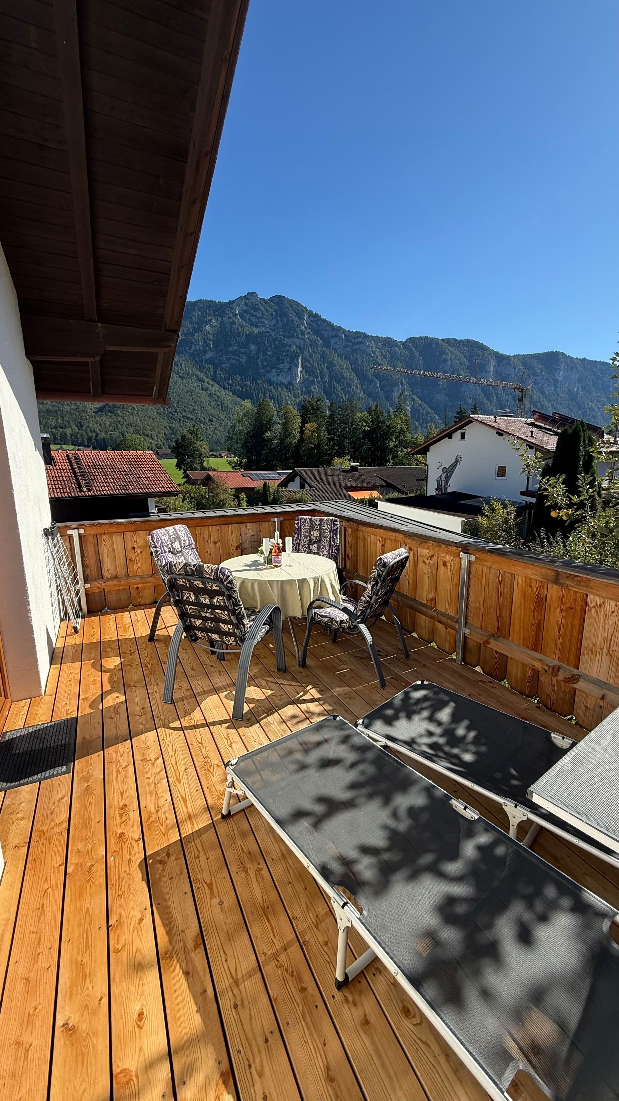 A wooden deck with a table and chairs, a sun lounger, and a view of mountains.