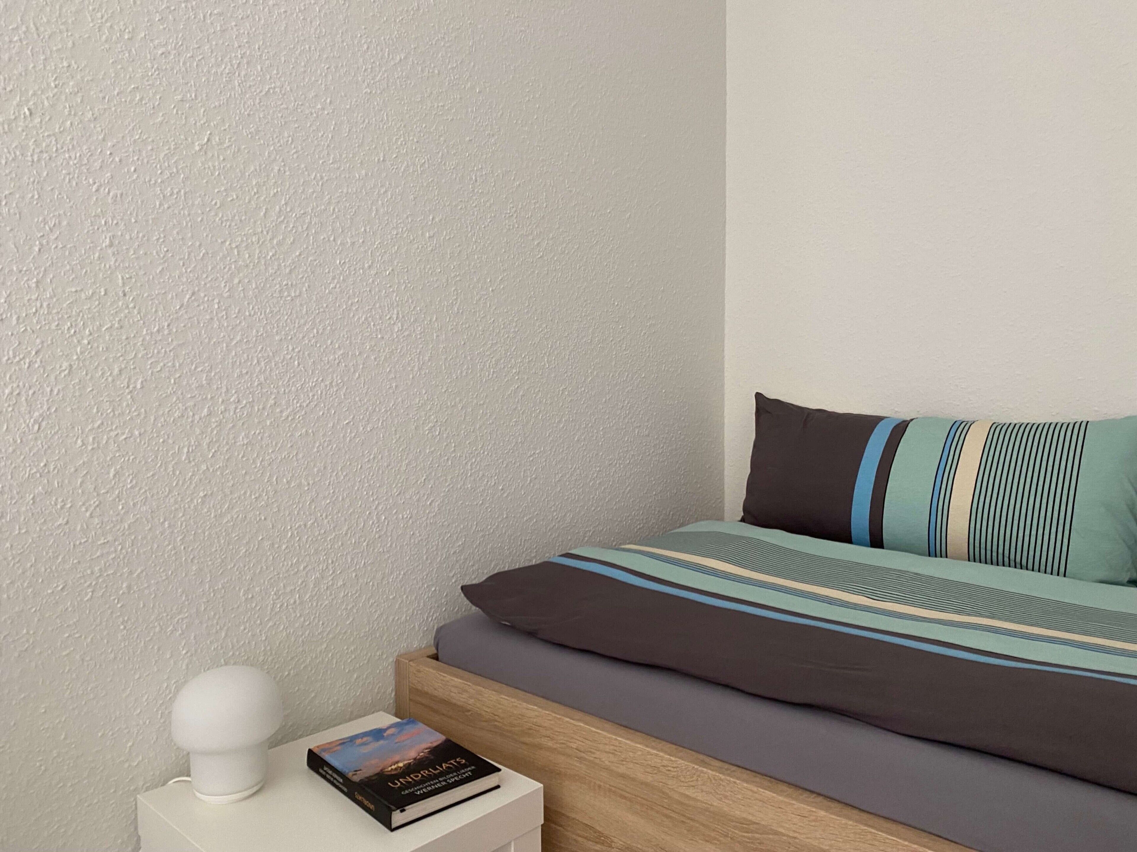 Ein ordentlich bezogenes Bett mit gestreifter Decke und Kissen, ein Nachttisch mit Buch und einem weißen Gegenstand, sowie eine strukturierte Wand.