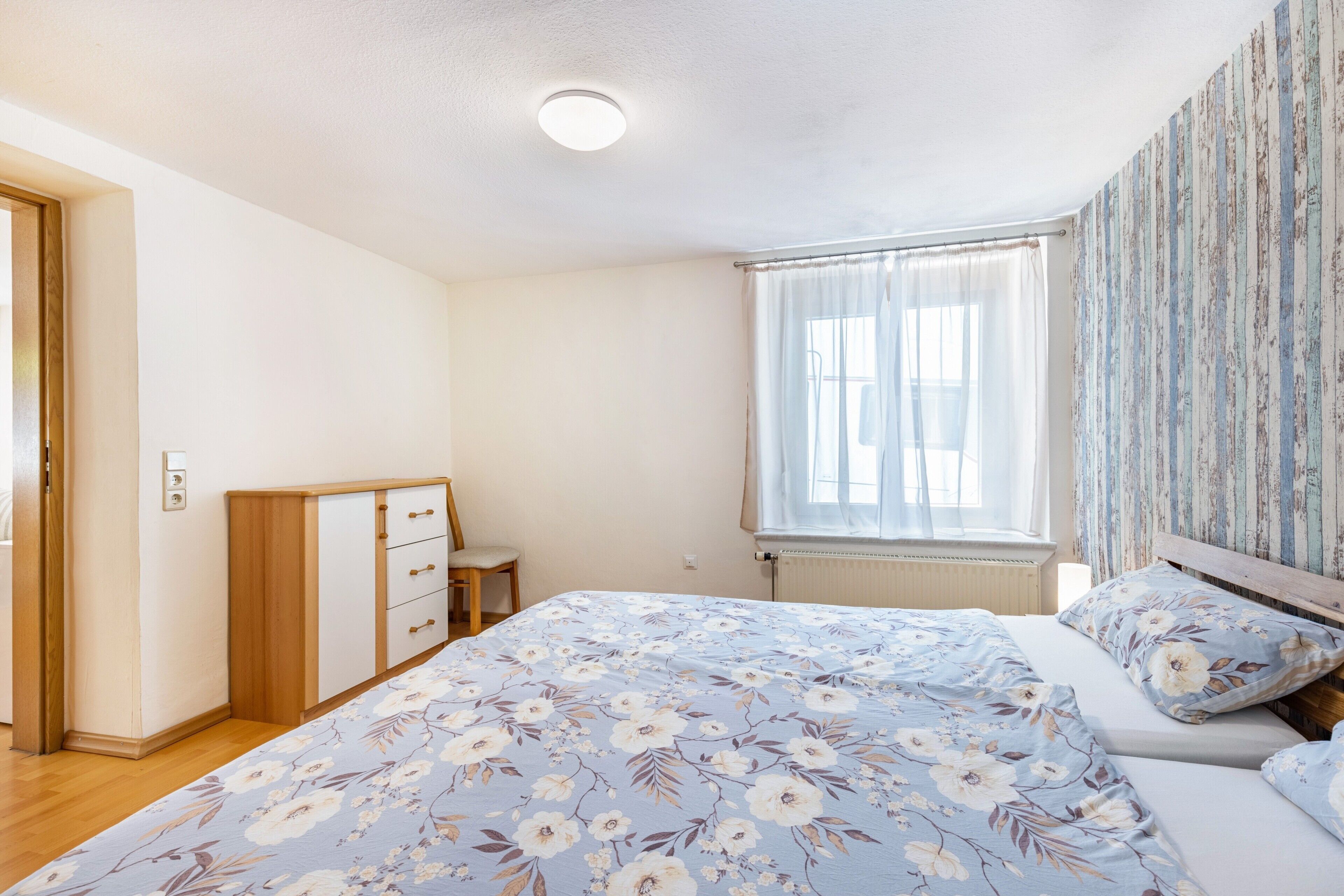 A bedroom with a bed, a dresser, a window with curtains, and a radiator.