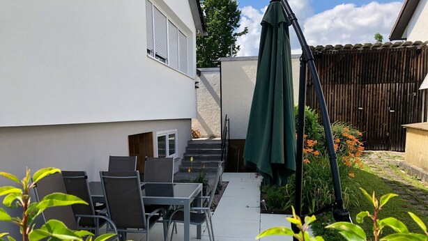Une terrasse avec des meubles d’extérieur, un parasol vert et une clôture en bois.