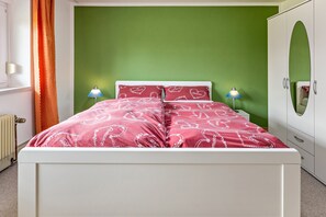 A bed with a red patterned bedspread, two bedside lamps, a white wardrobe with a mirror, and a green wall.