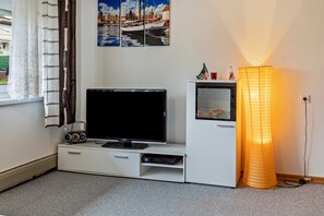 A room with a flat-screen TV, a small cabinet, a lamp, and a window with curtains.