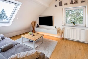Un salon moderne avec un plafond en pente, un téléviseur à écran plat fixé au mur, une table basse en bois, un canapé gris et une grande fenêtre donnant sur les arbres.