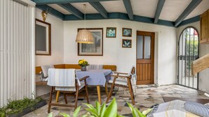 Um alpendre coberto com uma mesa, cadeiras, um banco de madeira, uma porta e uma janela.