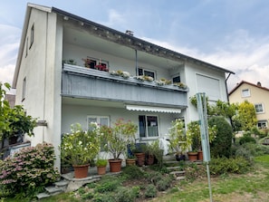 Un edificio de dos pisos con balcón, plantas en macetas y un área ajardinada.