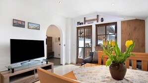 A living room with a flat-screen TV on a wooden stand, a dining table with a floral tablecloth, and a vase with yellow flowers. - Studio Apartment 'Lackner' with Mountain View, Balcony and Wi-Fi (Teisendorf)