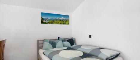 Un dormitorio con cama, una silla, un cuadro de un paisaje y una lámpara de techo.
