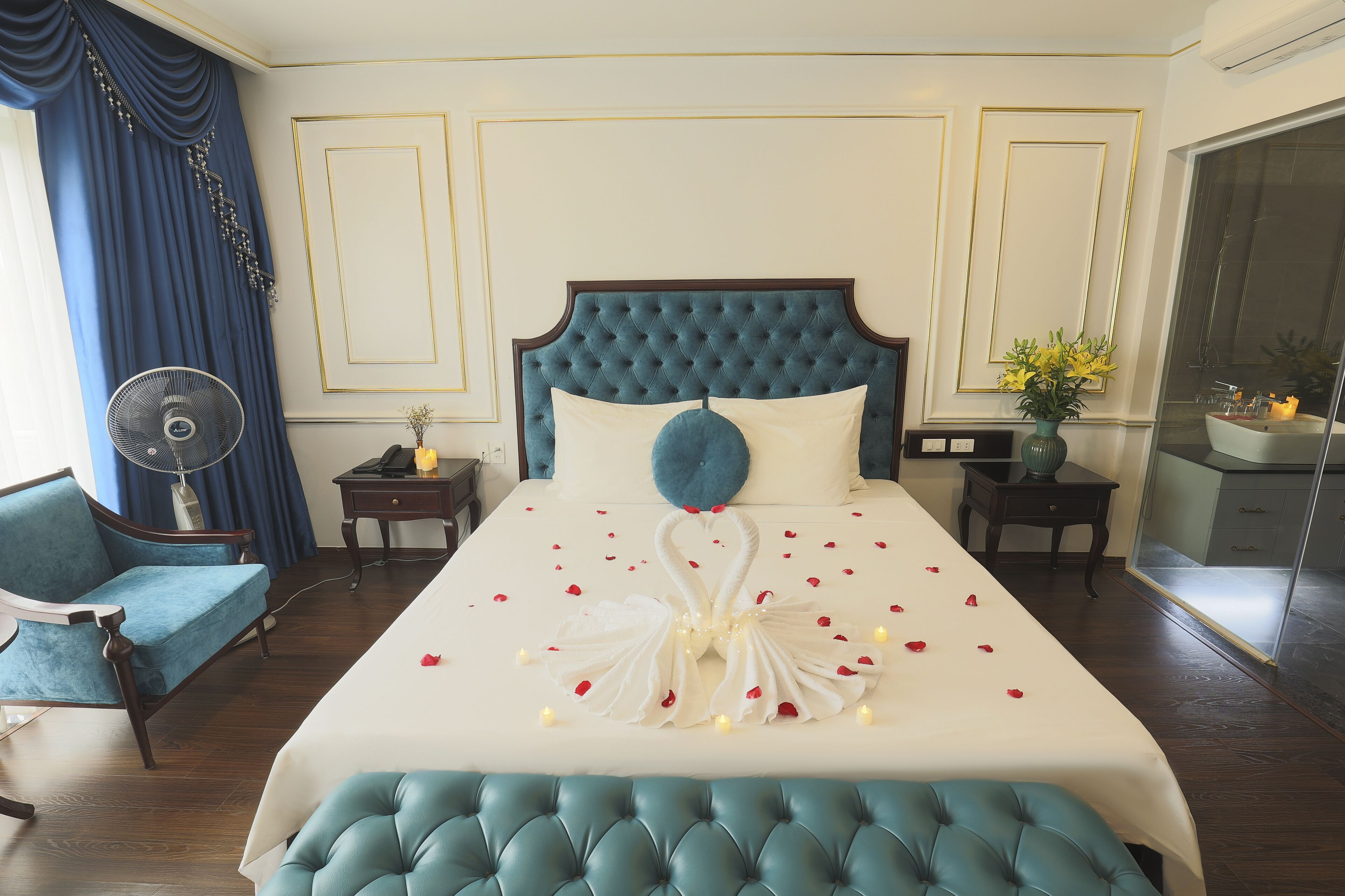 Habitación de hotel con una cama grande, un cabecero tapizado azul, un sofá azul, una mesita de noche con lámpara y un ventanal con cortinas.