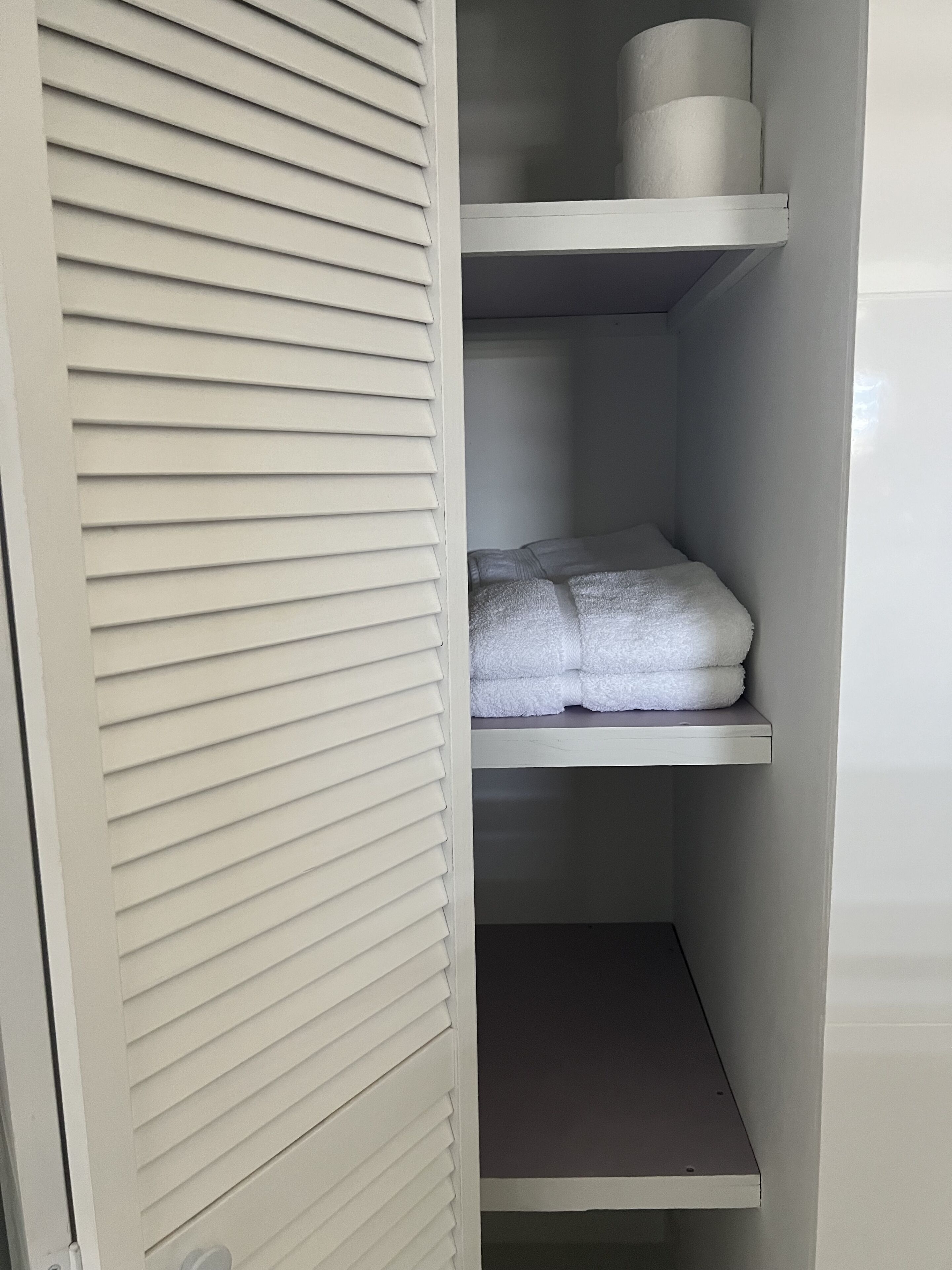 Um armário branco com venezianas horizontais, uma prateleira com toalhas dobradas e um rolo de papel higiênico na prateleira superior.