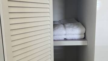 Um armário branco com venezianas horizontais, uma prateleira com toalhas dobradas e um rolo de papel higiênico na prateleira superior.