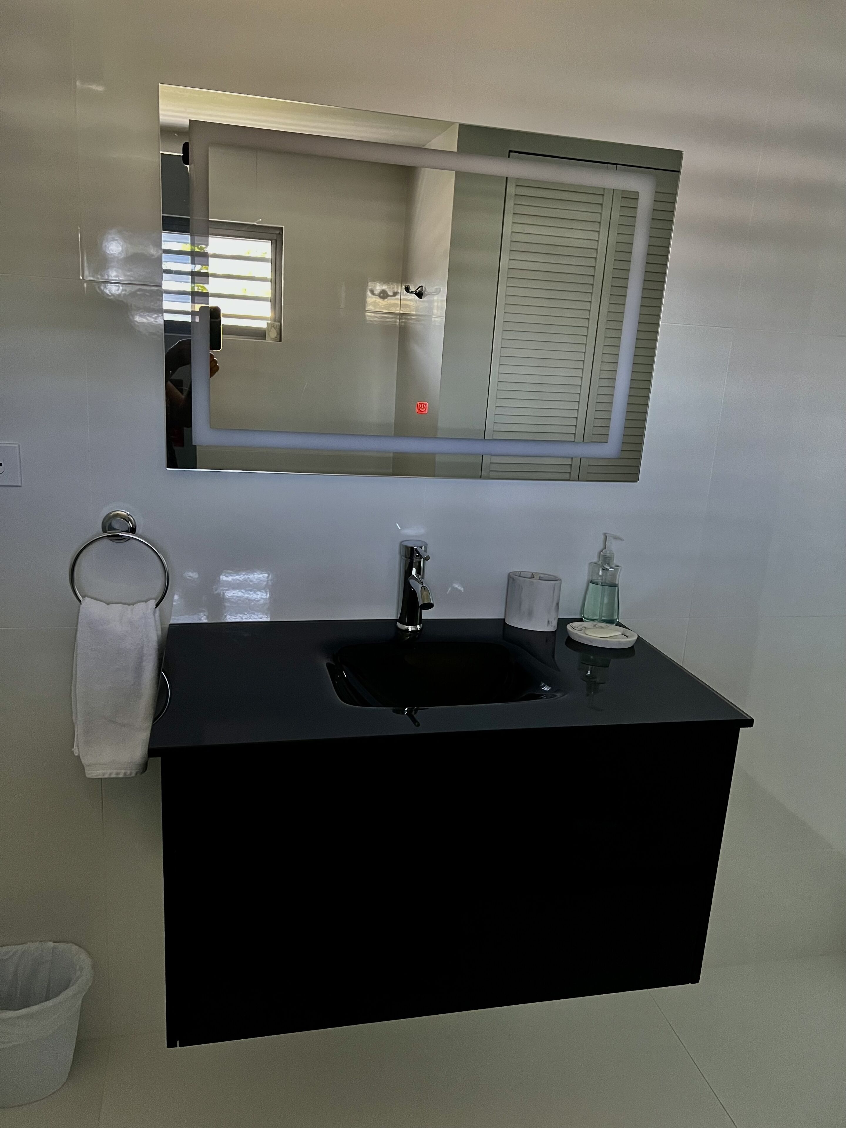 Un baño con un lavabo negro, un espejo, un perchero para toallas y un cubo de basura.