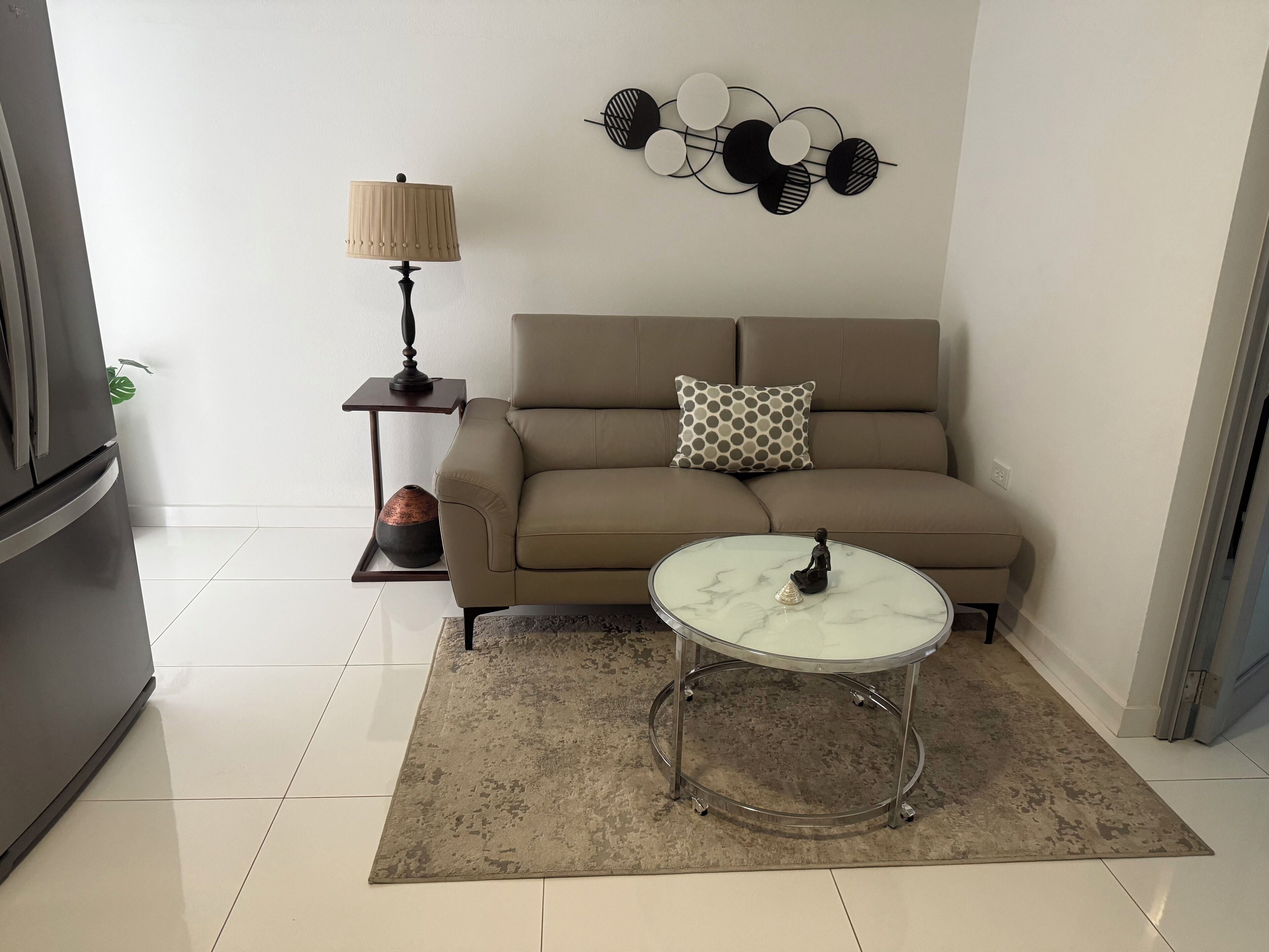Una sala de estar con un sofá color beige, una mesa redonda, una lámpara, un adorno de pared y una planta pequeña.