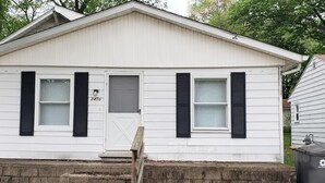 A single-story white house with black shutters, a front door, and a small set of stone steps leading up to the entrance.