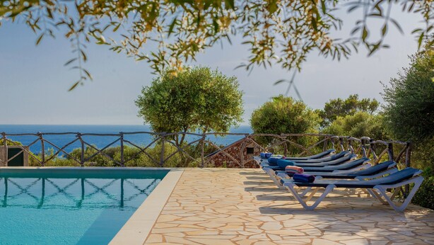 Uma piscina com um deck de pedra, cadeiras de descanso e vista para o mar.
