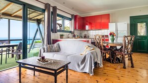 Un vaste espace de vie comprenant un canapé, une table à manger et des chaises, une cuisine avec des meubles rouges et une grande fenêtre donnant sur l’océan.