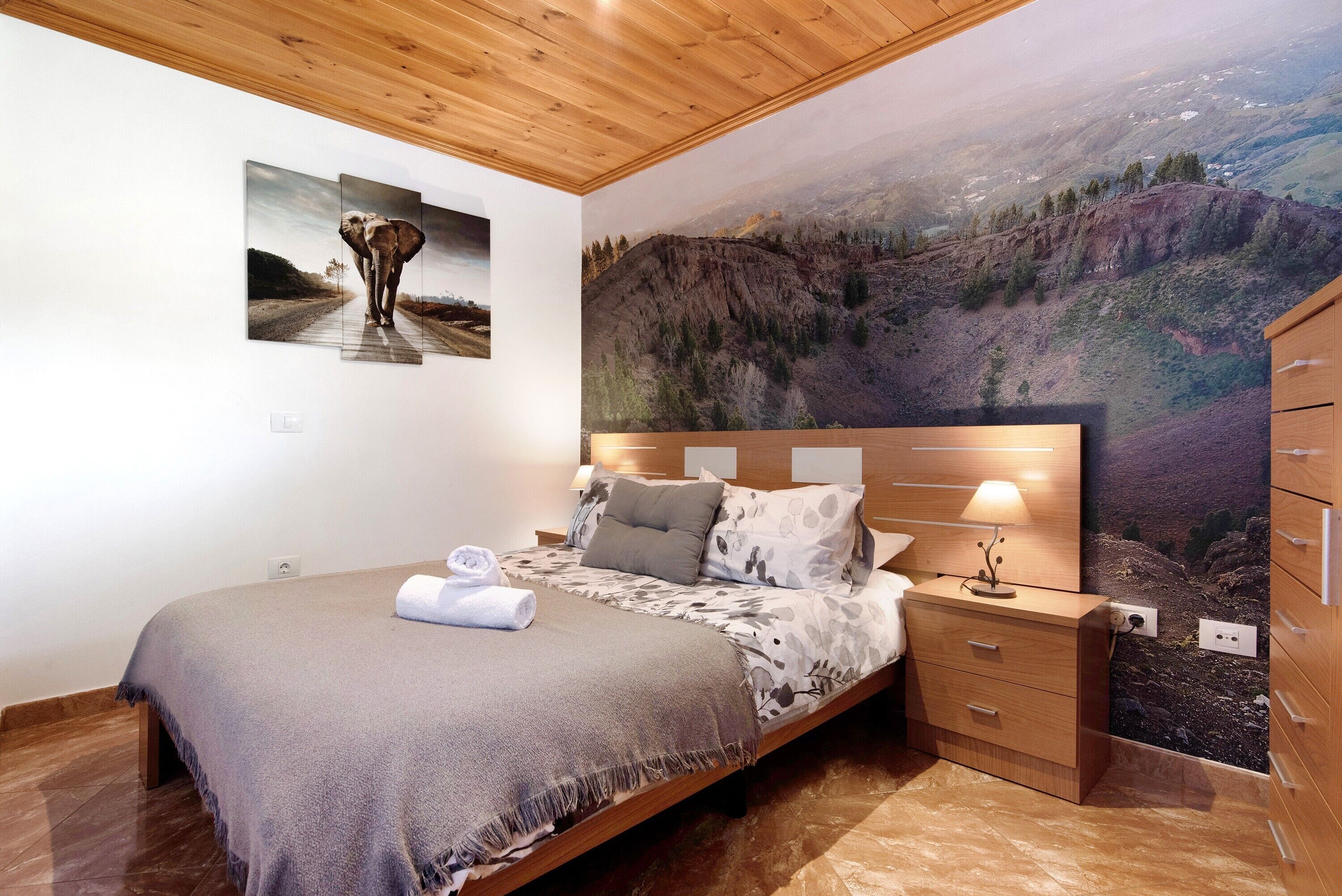 Un dormitorio con una cama, una mesita de noche, un aparador y un amplio mural de un paisaje montañoso.