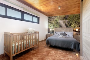 A bedroom with a crib, a bed with a blanket, a nightstand with a lamp, and a view of a waterfall.