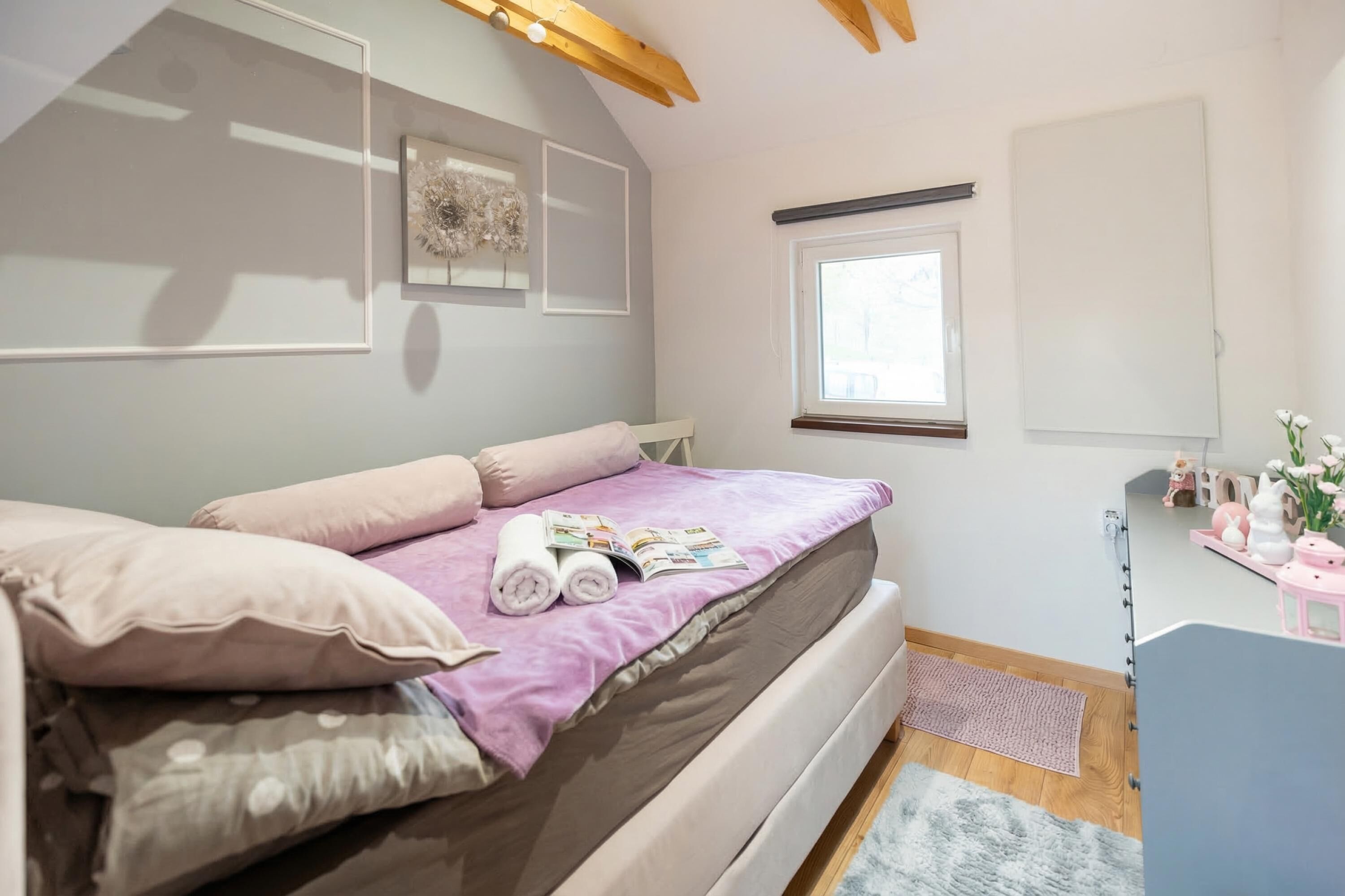 Un dormitorio con cama, almohadas, ventana, mesita de noche con lámpara y estante con adornos.