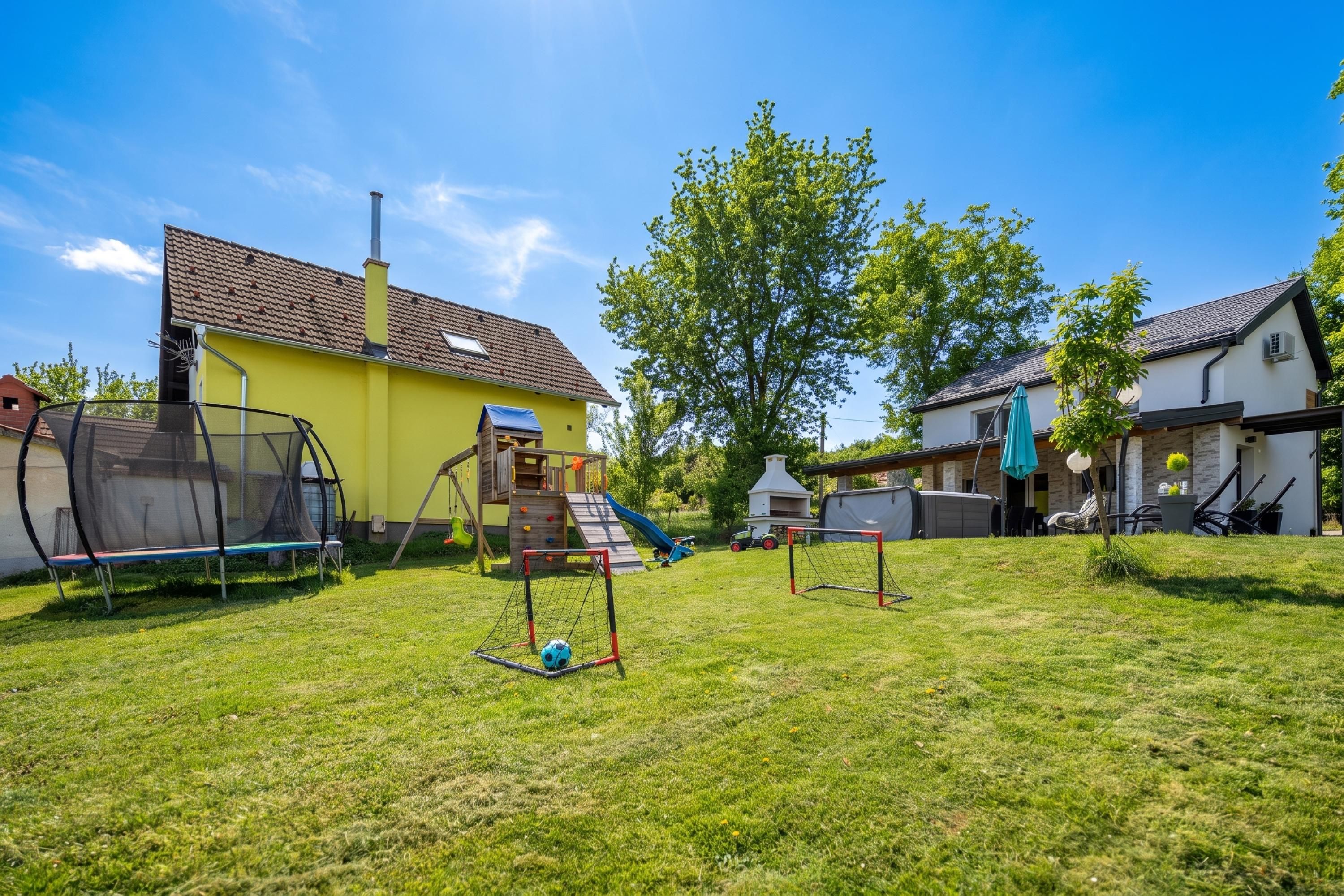 Een achtertuin met een trampoline, een speelhuisje en een voetbalkooi.