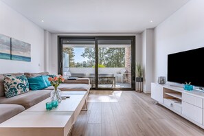 A modern living room with a sofa, coffee table, TV, and a sliding glass door leading to an outdoor area. - 206 Navi Golf – Family-Friendly Retreat with Pool & Kids Park (La Cala de Mijas)