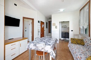 A room with a dining table, chairs, a sofa, a TV, and a kitchenette.