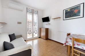 A living room with a sofa, a dining table with chairs, a television mounted on the wall, and a window with curtains.