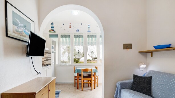 A living room with a TV, a dining table with chairs, a sofa, a window with a view of boats, and a small shelf with a bowl. - Studio Apartment "Monolocale 12" with Sea View, Wi-Fi & Air Conditioning (Pietra Ligure)