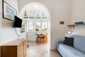 A living room with a TV, a dining table with chairs, a sofa, a window with a view of boats, and a small shelf with a bowl.